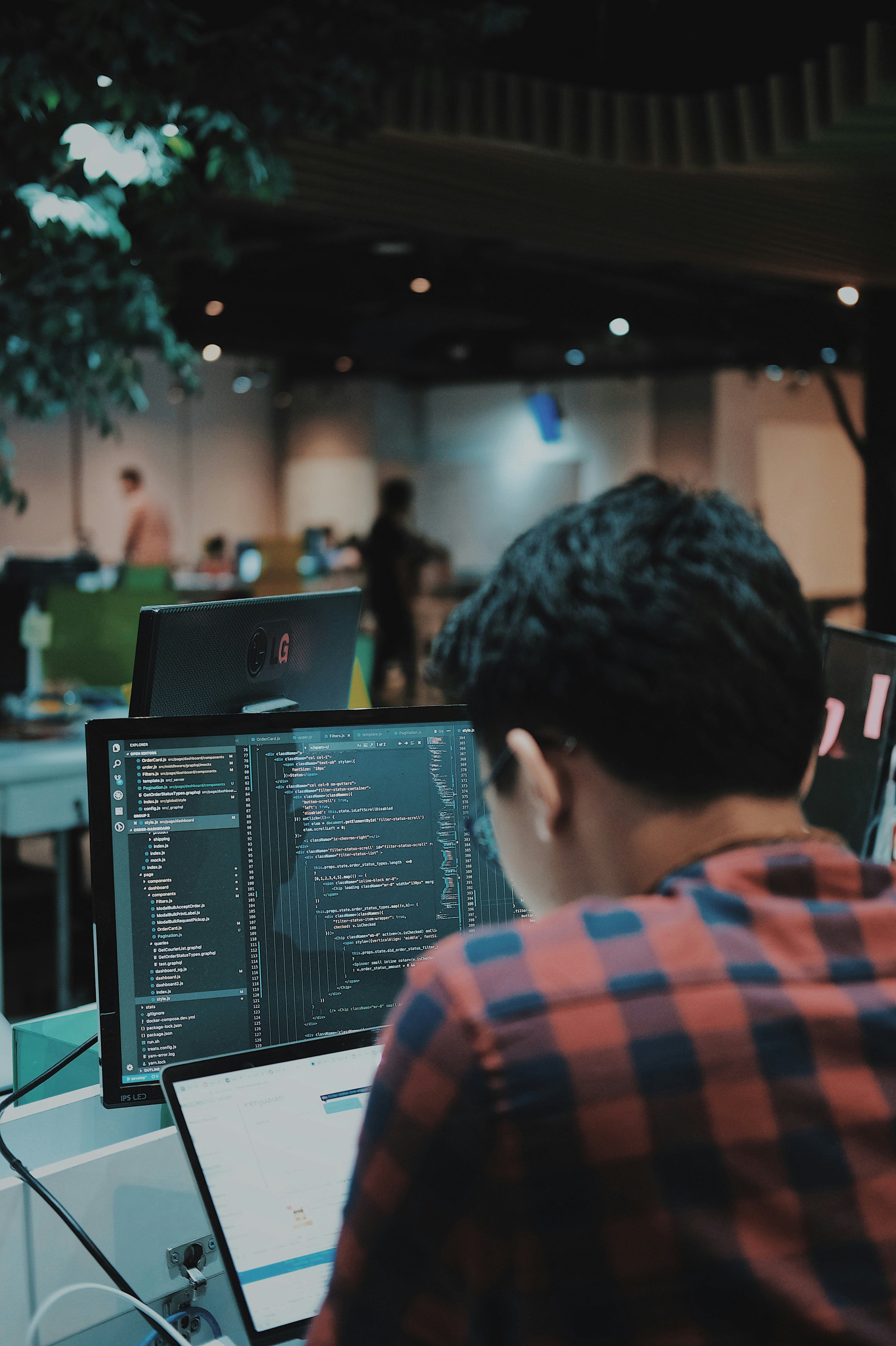 Developer in a plaid shirt reviewing code on a monitor in an open-plan office