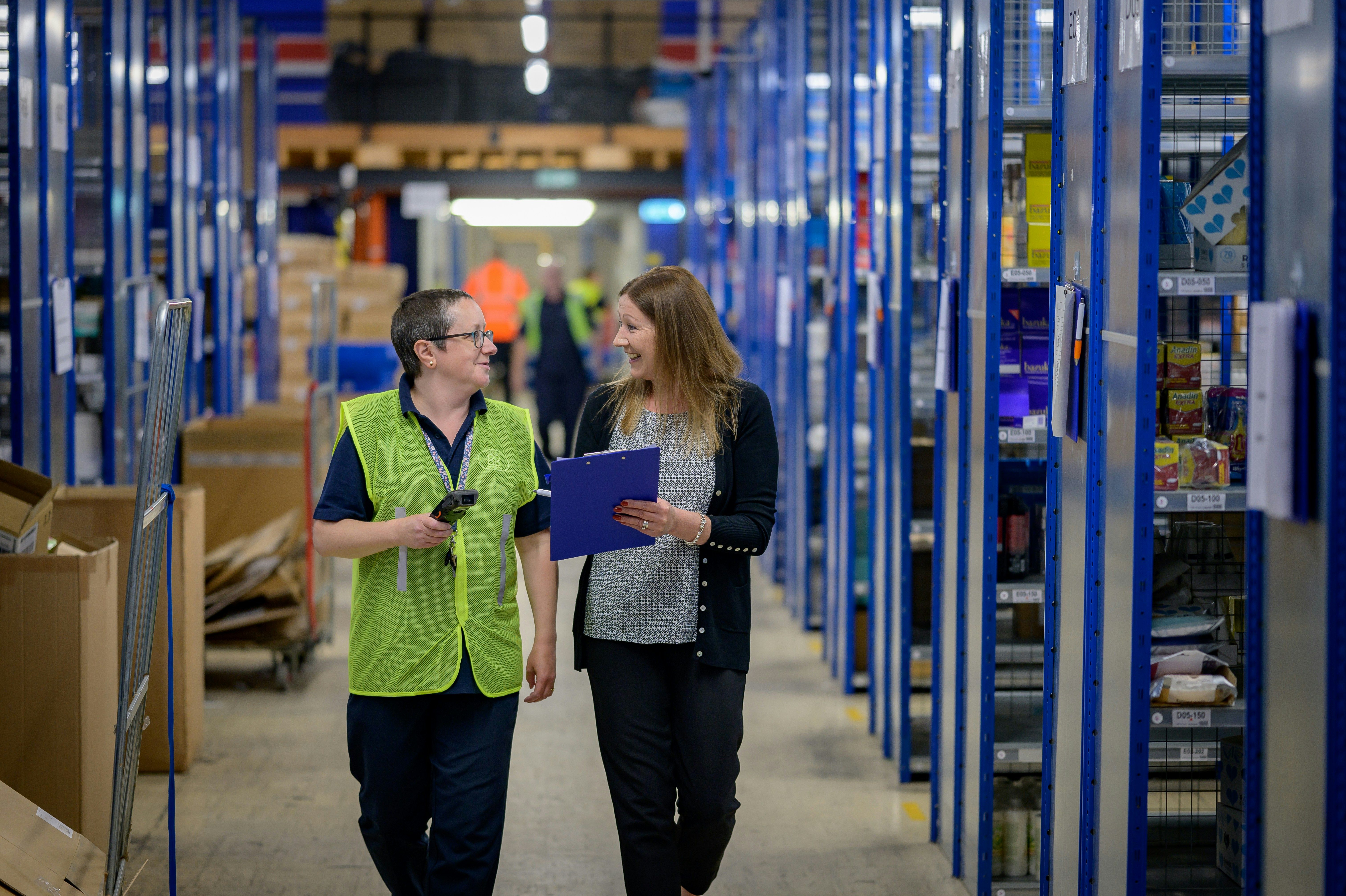 Two colleagues in a warehouse aisle between blue shelves, one holding a handheld device and one a clipboard
