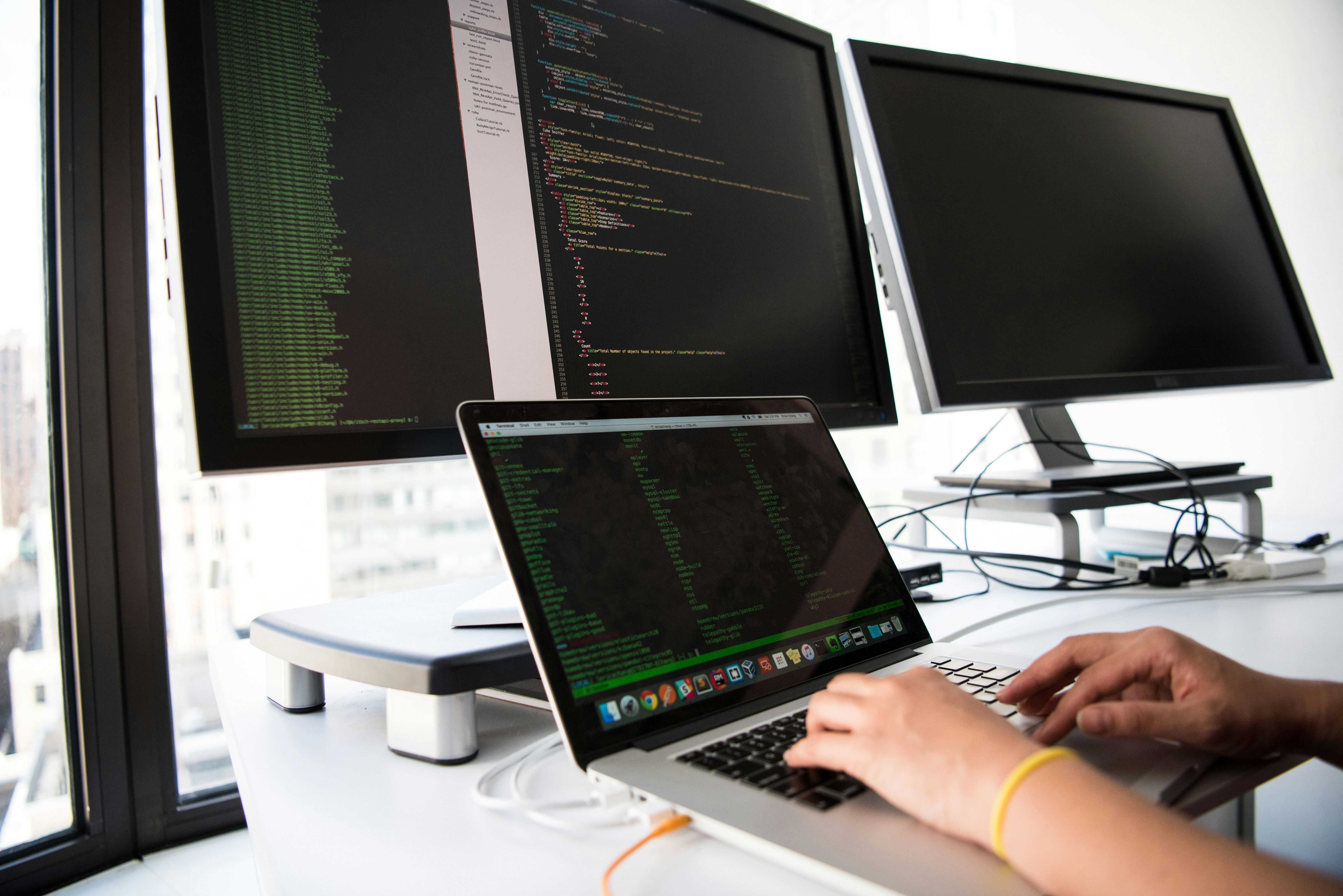 Hands typing on a laptop in front of monitors showing lines of code