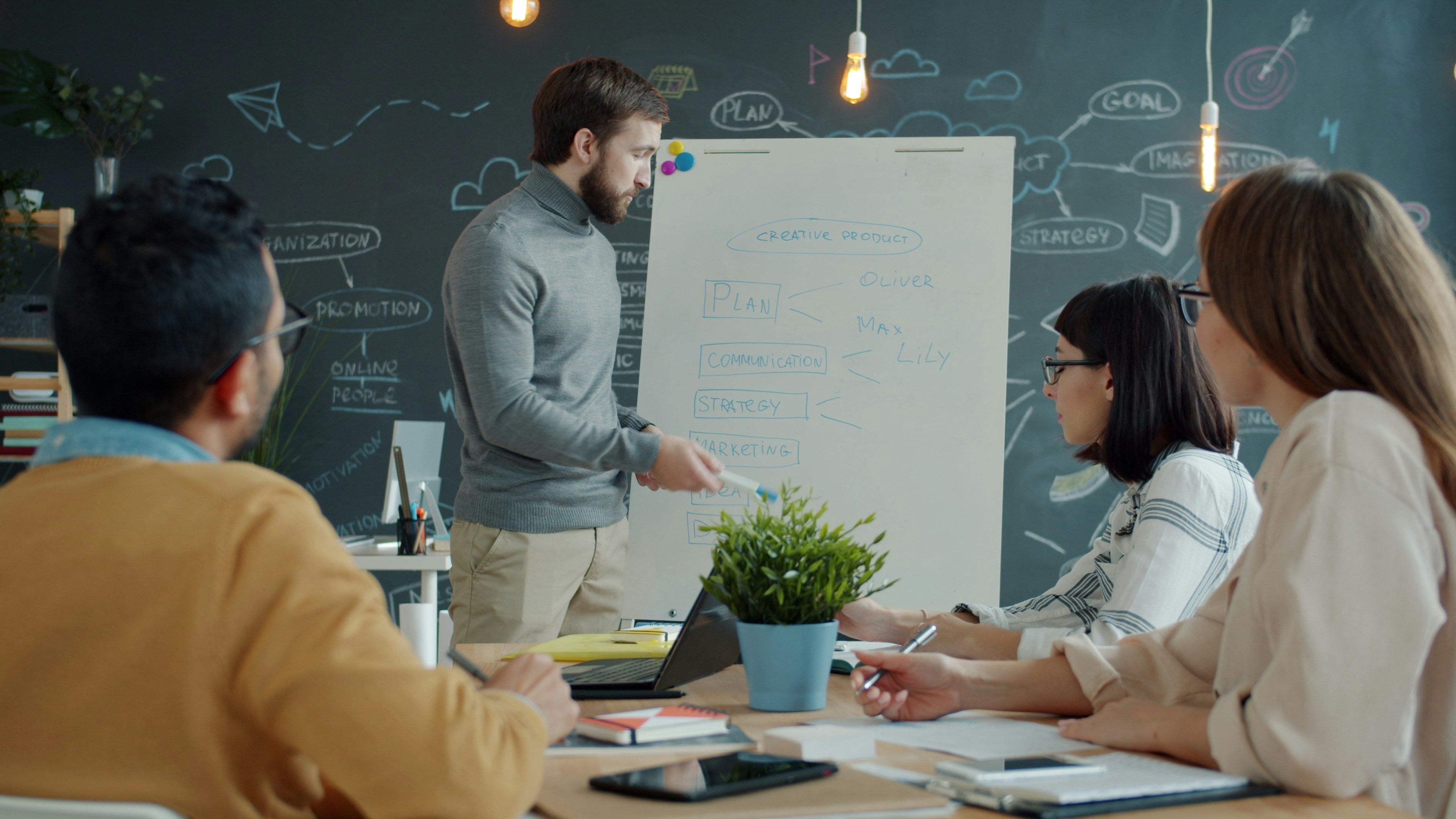 Colleagues around a table as a man presents a plan on a whiteboard